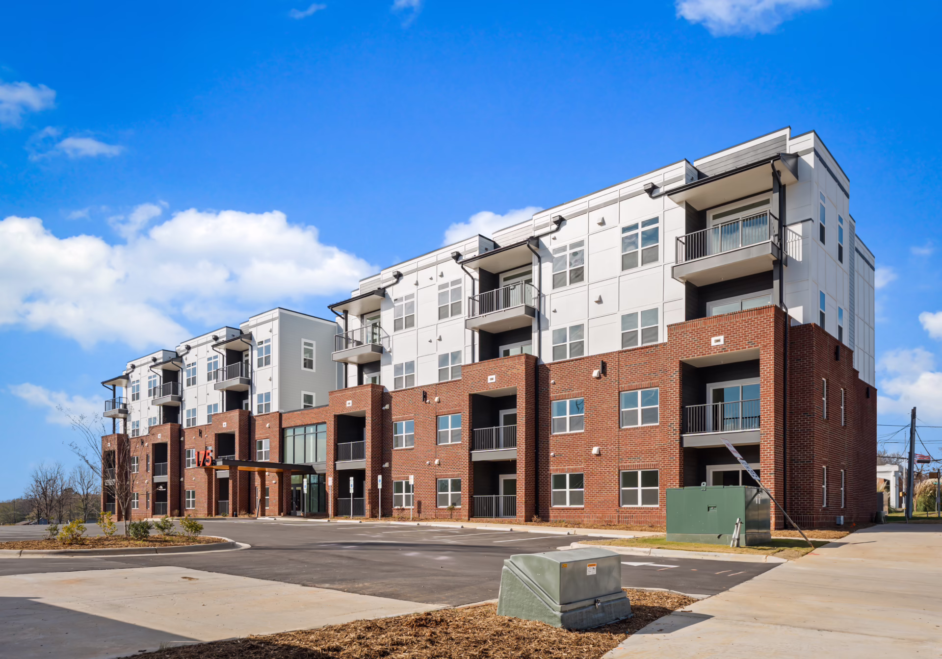 Brixton Apartments seen from the north corner: four storeys of red brick with pale metal panel above, recessed black steel balconies, and an open blue sky.