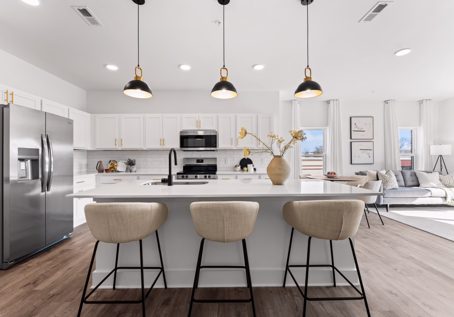 An apartment kitchen with a white quartz island and three upholstered barstools, white shaker cabinets with brass pulls, stainless appliances, and three matte-black pendant lights overhead.