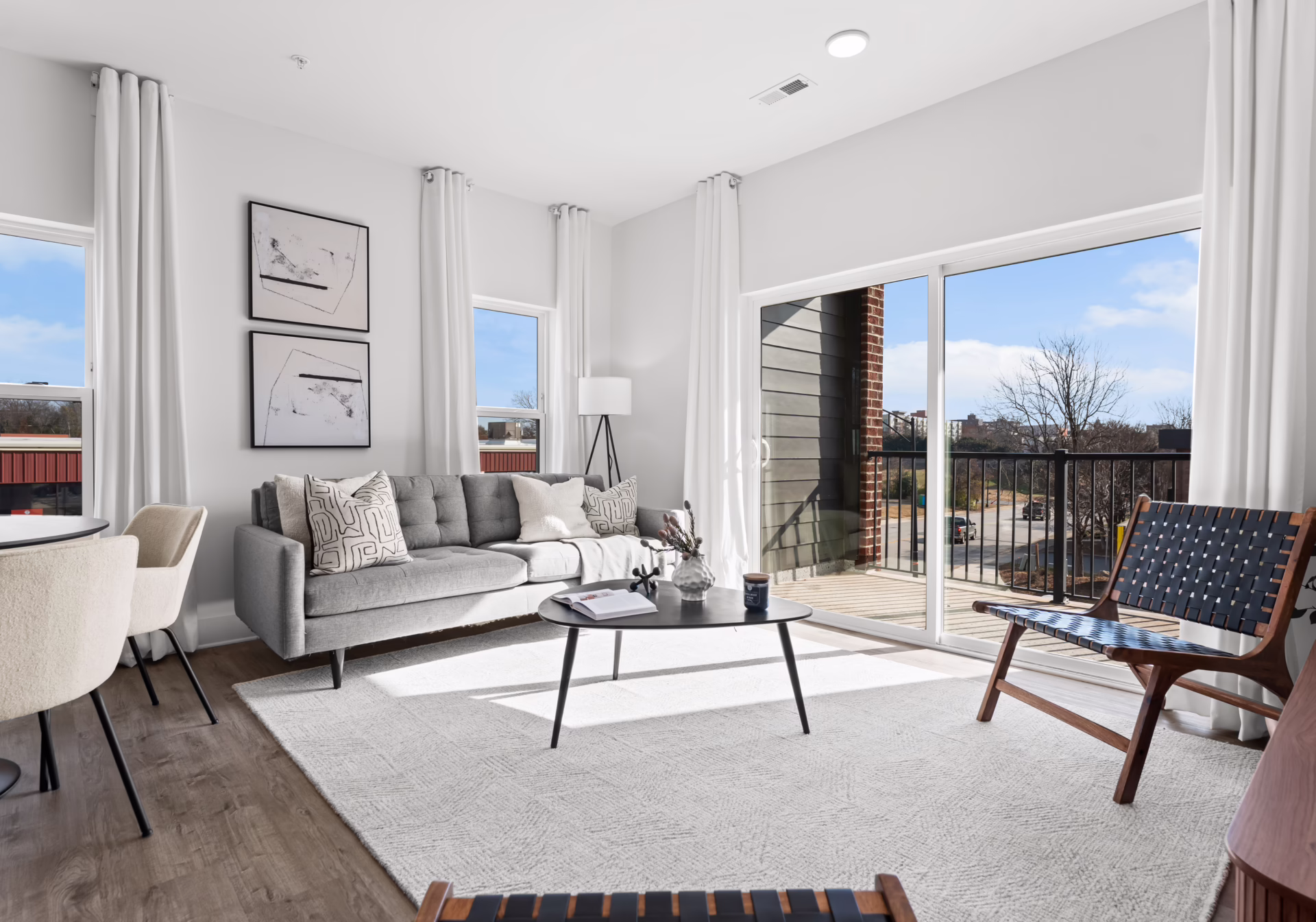 An apartment living room with a grey sofa, patterned cushions and a sliding glass door opening onto a balcony.